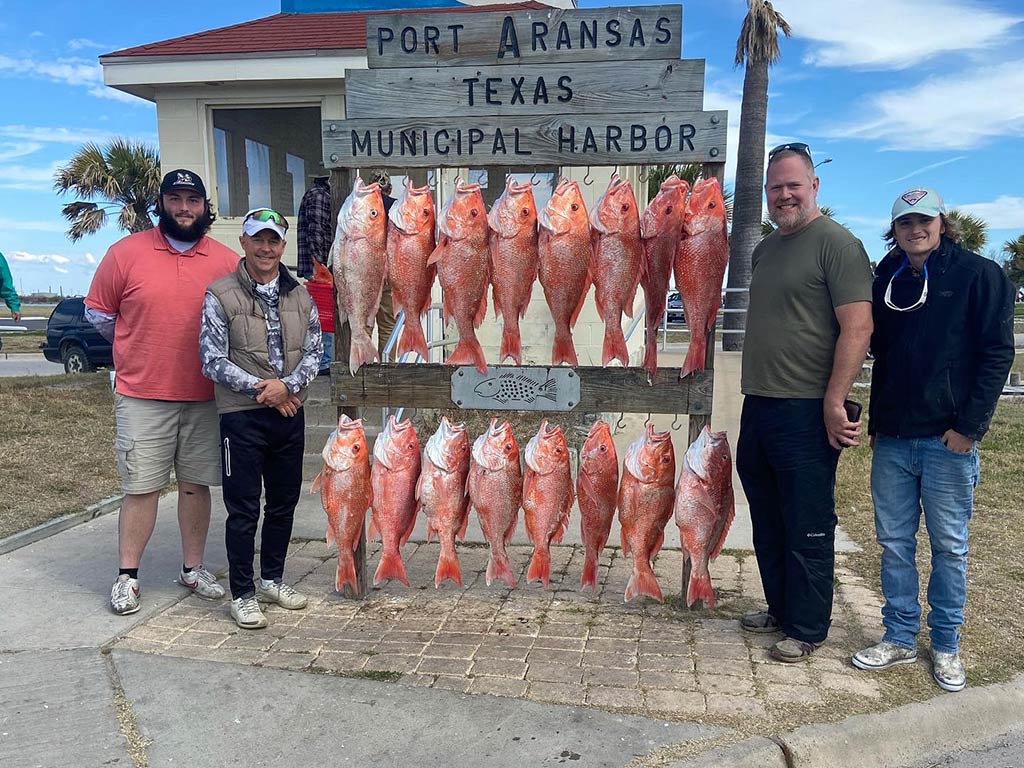 Four anglers pose next to a wooden sign that says "Port Aransas, Texas, Municipal Harbor", with over a dozen Red Snappers hanging from hooks on it