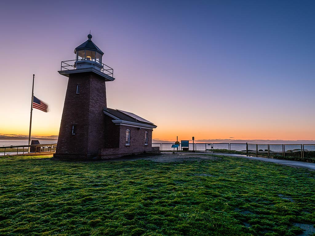 A view towards the Santa Cruz lighthouse at sunset, with a US flag at half mast to the left of it, with the Pacific Ocean in the background