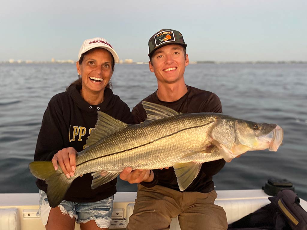 A male and female angler sat on the side of a boat near sunset, holding a large Snook with the water behind them