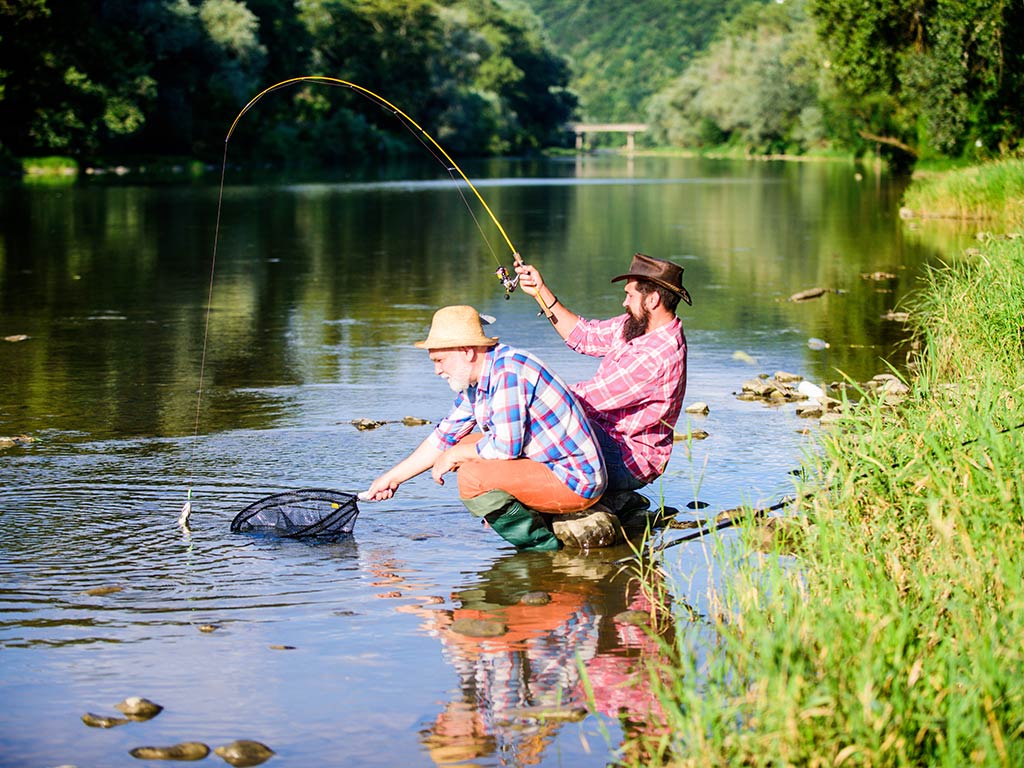 An elderly and middle-aged angler crouching in the waters of a river on a sunny day, while the younger man holds a fishing rod and the elder is netting a fish