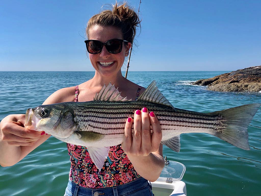 A female angler wearing sunglasses, holding a Striped Bass while standing on a boat with the water and a rocky shoreline behind her on a sunny day