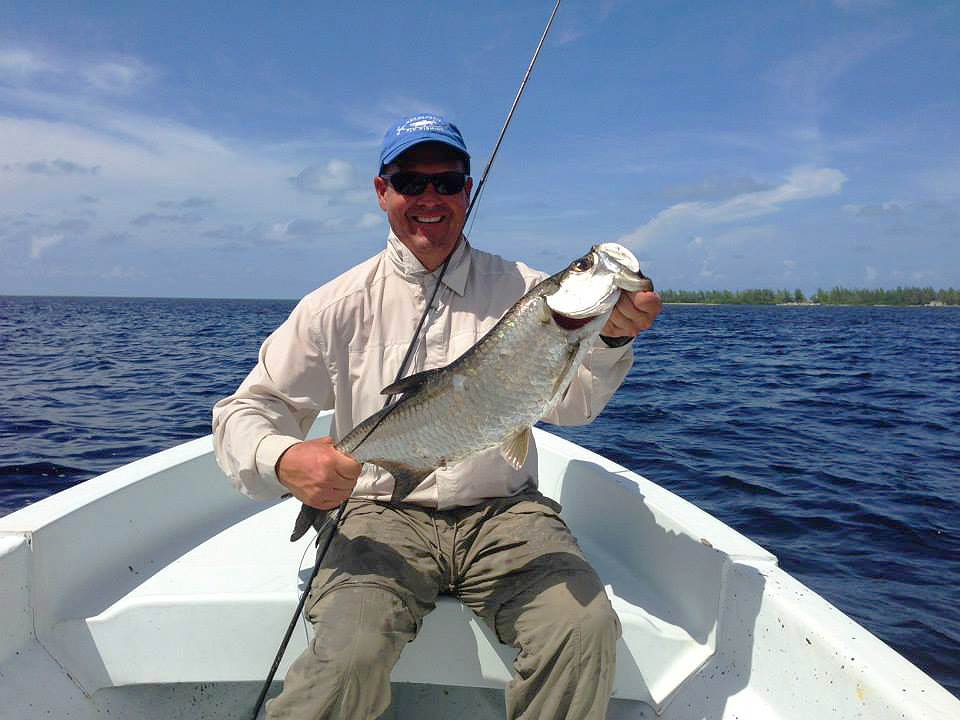 A smiling middle-aged angler holding a Tarpon with a fly fishing rod leaning next to him on a boat on the water in Mexico