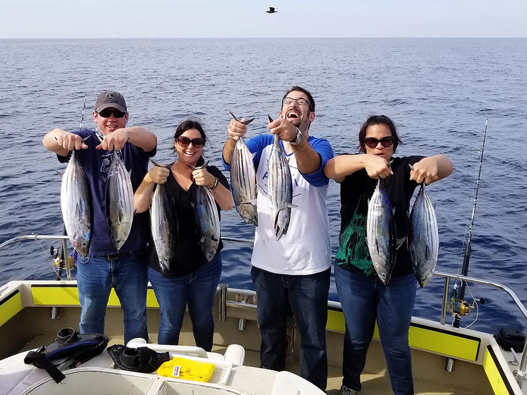 Four anglers, two male and two female, holding two Tunas each at the back of a fishing boat, with the water behind them and a bird flying above them in the distance