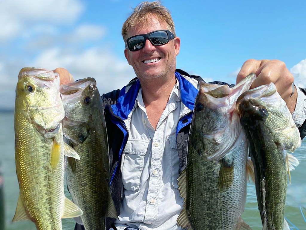 A smiling angler wearing sunglasses, holding four Bass caught while fishing in Lake Travis on a sunny day