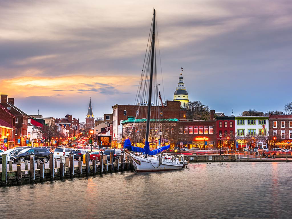 A view across the water towards downtown Annapolis, with the Maryland state capitol building visible in the distance behind a boat and some buildings on a cloudy evening