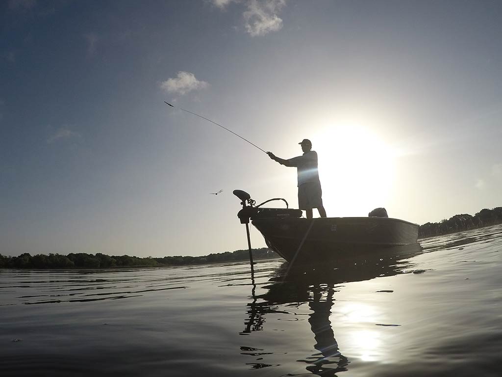 A view across the water of an angler casting a line from the front of a boat in Lake Travis on a sunny day, with the sun behind them in the distance