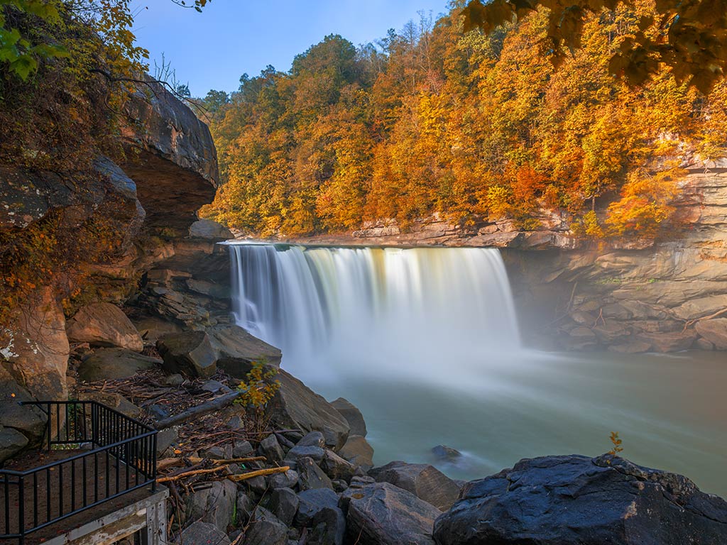 A view of Cumberland Falls near Cumberland Lake in the Kentucky Wetlands, with fall foliage surrounding the water on a clear, sunny day