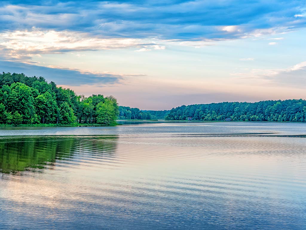A view across the calm water of Lake Chesdin near sunset in Virginia, with foliage visible around the lake and clouds in the sky