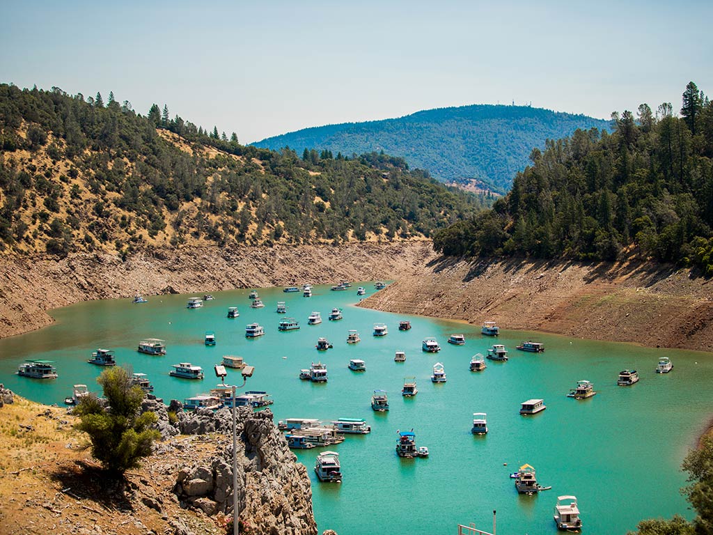 A view from a hill of a well-organised line-up of boats in a harbor in Lake Oroville, California, with turquoise waters visible and typical Californian hills in the distance