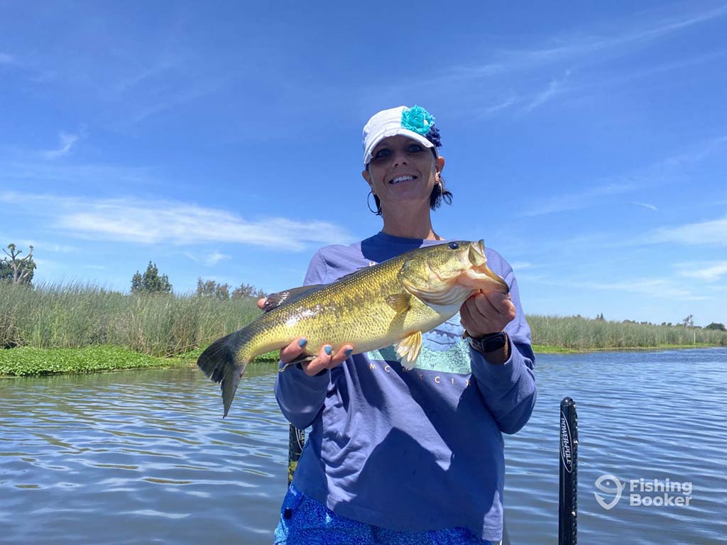 A smiling female angler in a baseball cap, stands on a dock with the water and some foliage behind her on a sunny day, showing off her Largemouth Bass catch