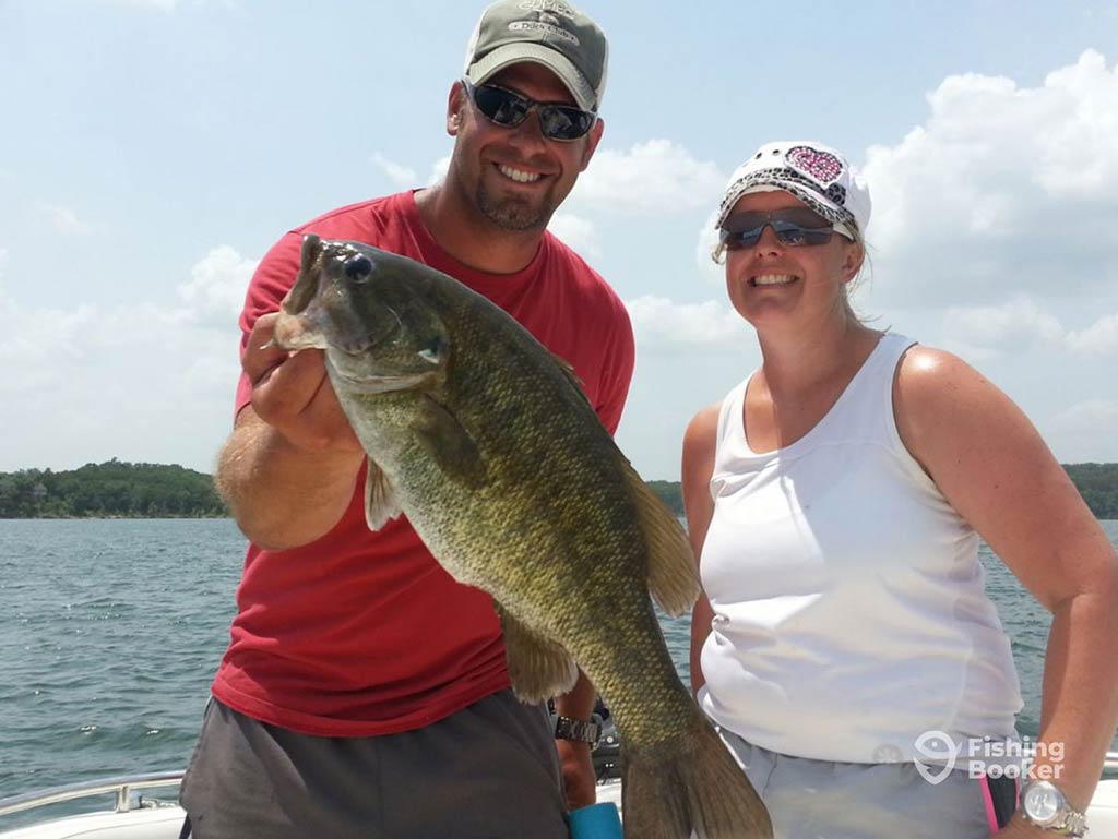 A man in sunglasses holds up a Largemouth Bass to the camera, while a woman in a white vest next to him smiles on a sunny day, with water behind them and a green shoreline in the distance