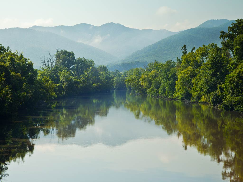 A view across the calm waters of the Nolichucky River in Tennessee, with a large mountain visible in the distance and green trees surrounding the water on a hazy, sunny day