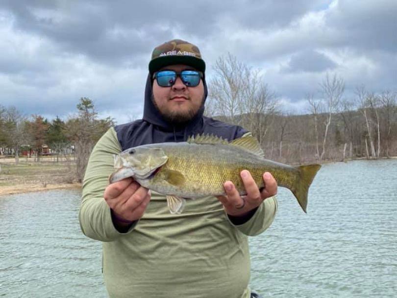 A man in a baseball cap and sunglasses, holding a Smallmouth Bass to the camera on Table Rock Lake, with the water and fall foliage behind him on a cloudy day