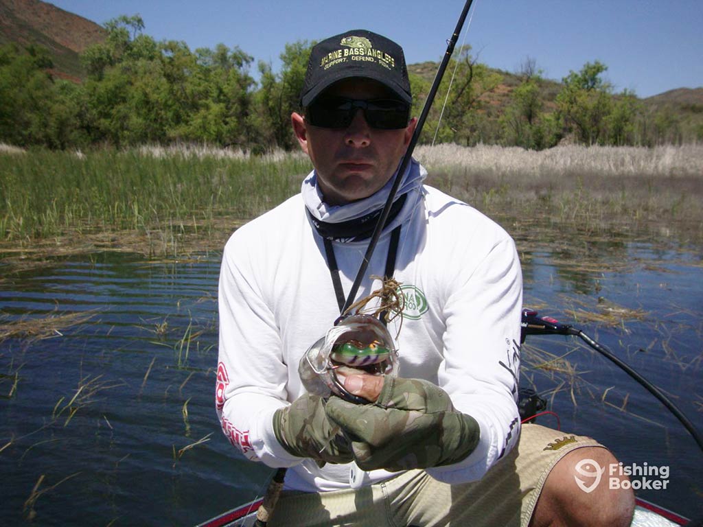 An angler wearing sunglasses and a baseball cap crouches down on a boat with a rod across his front, as he presents a Largemouth Bass caught on artificial bait to the camera