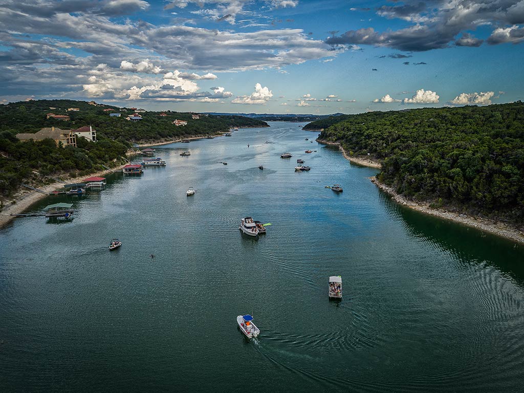 An aerial view of a busy waterway on Lake Travis, Texas, with boats filling the water and land on either side on a day with sunny intervals