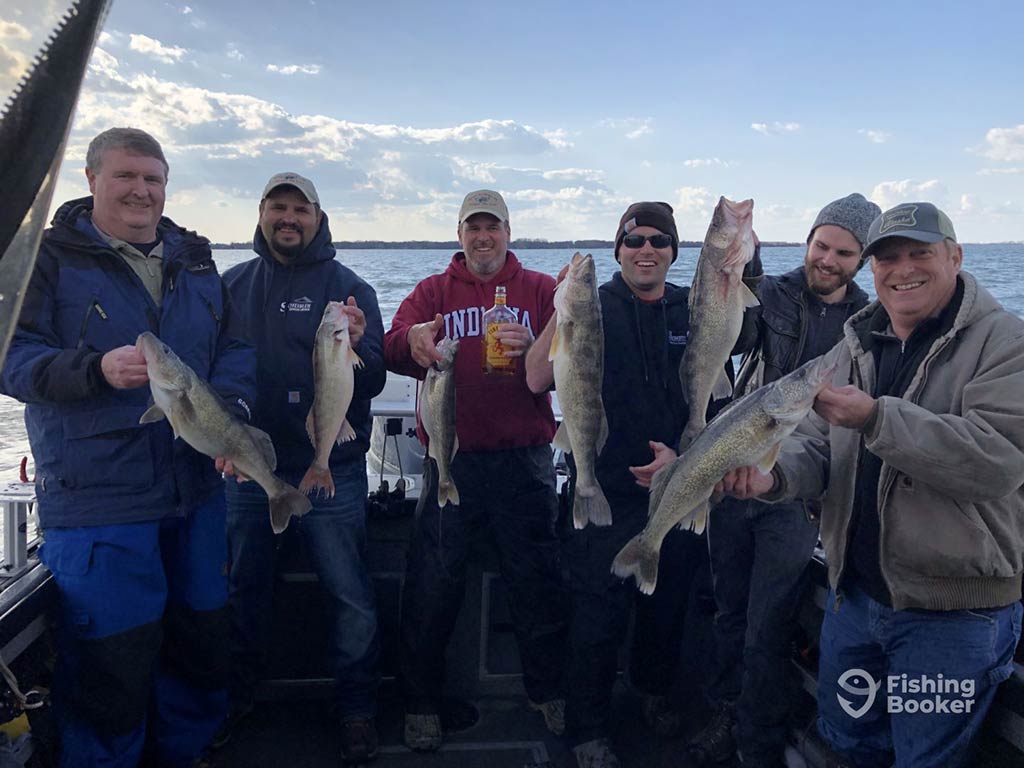 A group of male anglers stood on the deck of a fishing boat, holding a load of Walleye they caught, with the water behind them at the end of a clear day