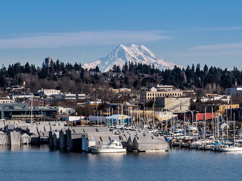 A view across the water towards downtown Olympia, Washington, with a snow-capped mountain in the distance on a sunny day, featuring boats in the harbor in the foreground