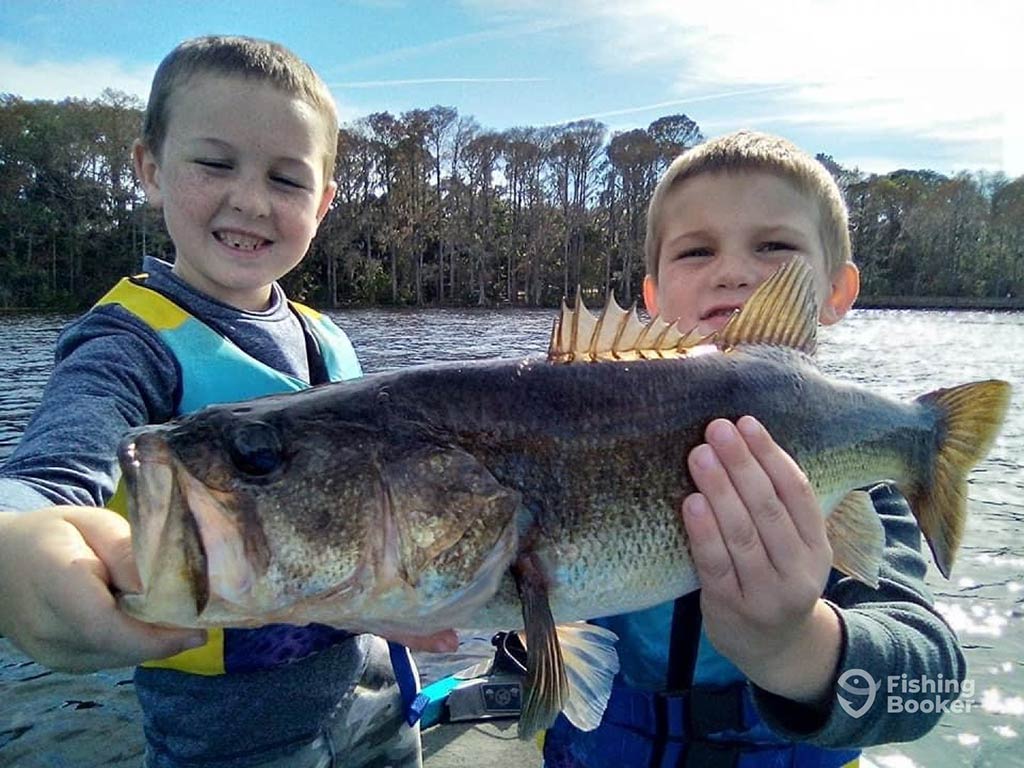 Two children holding up a Bass to the camera, while smiling, standing on a boat with some trees on the shoreline behind them on a sunny morning