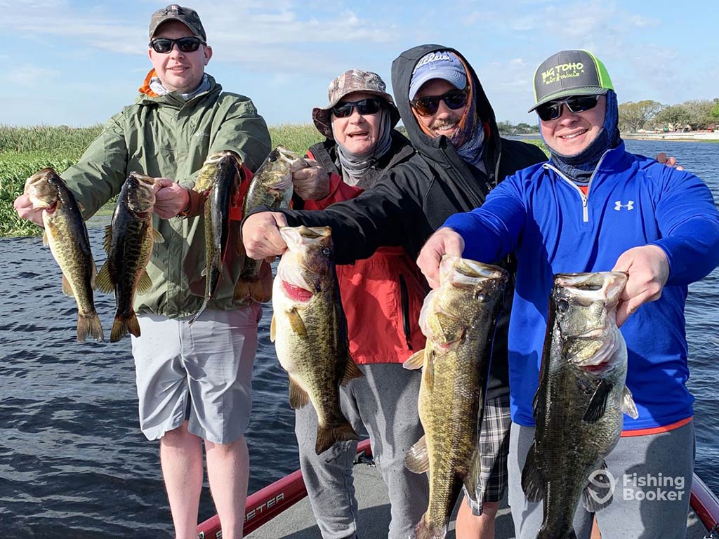 A group of four male anglers standing on a boat, holding up a Bass each, while wearing caps, sunglasses, and raincoats on a day with sunny intervals