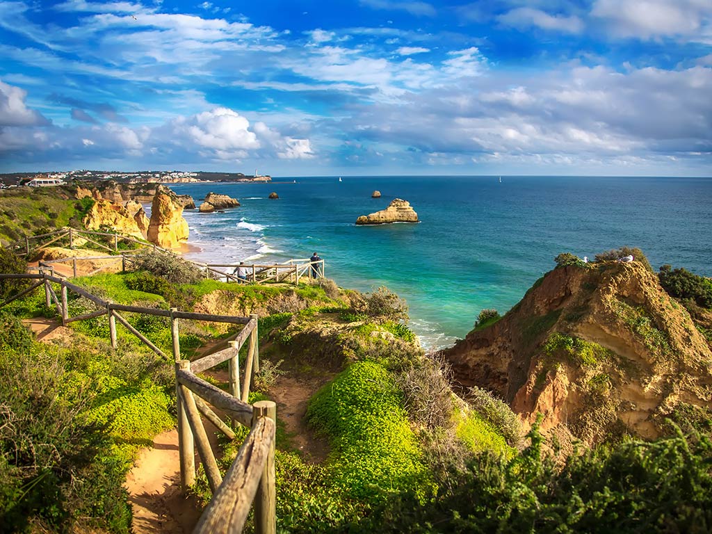 A view from a path on hill towards the rugged, rocky coastline of the Algarve, with crystal clear waters visible in the distance on a sunny day