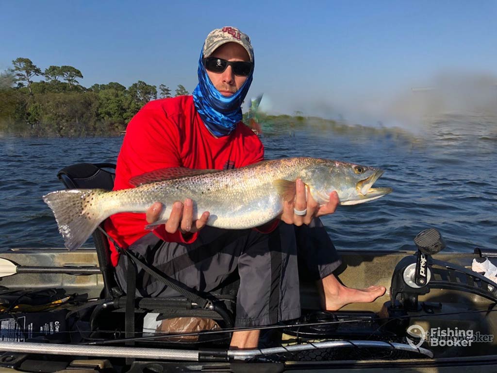 An angler in a baseball cap, sunglasses, and neck bluff, sitting on a kayak and presenting a Spotted Seatrout to the camera on a sunny day