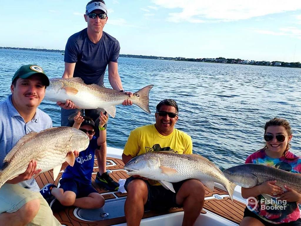 A group of three anglers sit on a boat, while one other angler stands behind them, holding up a large Redfish each, with the water behind them on a sunny day