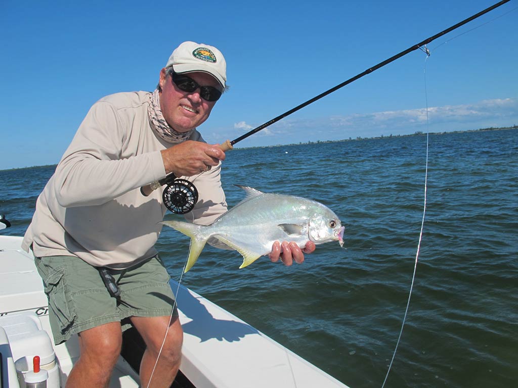 A man posing on the side of a boat, while holding a 4 lb pound Pompano, over a shallow shoal in the Indian River Lagoon near Jensen Beach, Florida on a sunny day