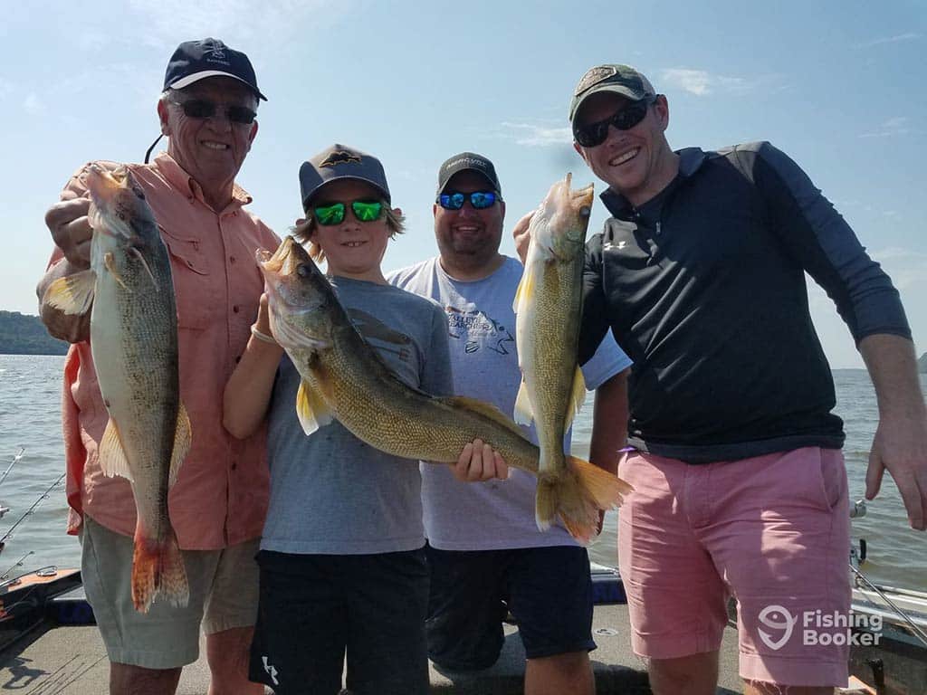 A group of four anglers of different ages, standing on a fishing boat and holding three Walleyes caught while fishing on the Mississippi River on a sunny day