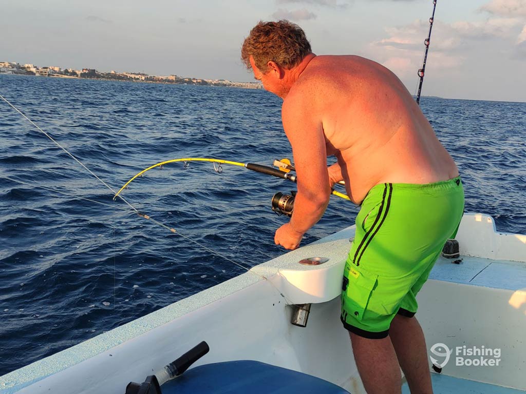 A shirtless angler bends over the side of the boat, holding a fishing rod in his hands, as he battles a fish in Puerto Morelos near sunset