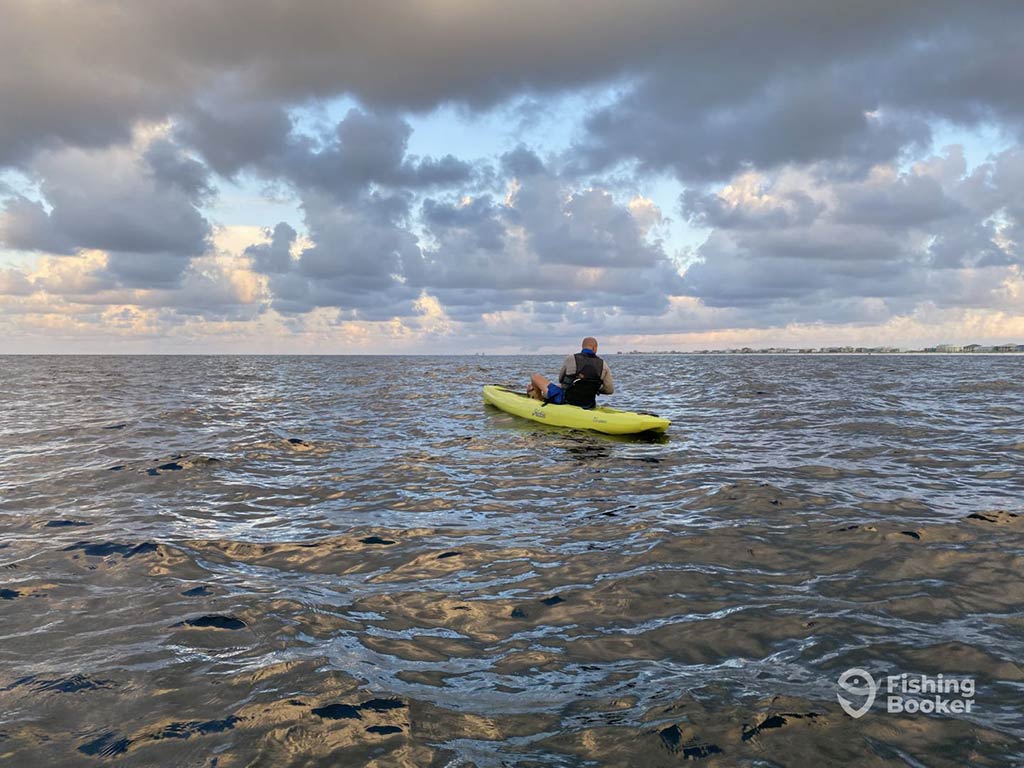 A view across the water of a lone kayak angler heading out along the calm waters of Navarre, FL, on a cloudy day