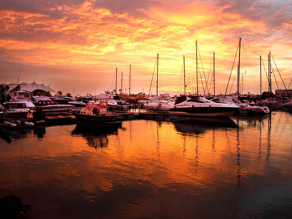 A view across the water towards a busy marina full of boats at sunset in the Algarve, with clouds in the orange sky