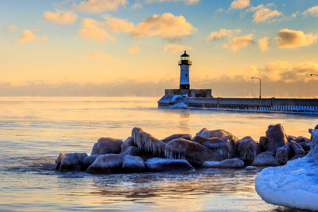 A view of the Duluth Lighthouse at sunset, with some rocks in the water and remnants of snow on them