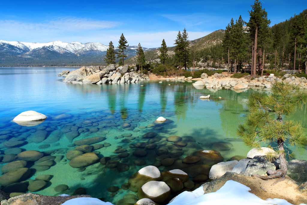 Crystal clear and blue waters of Lake Tahoe, with woods on the shore and clear blue skies, along with snow-capped mountains in the distance