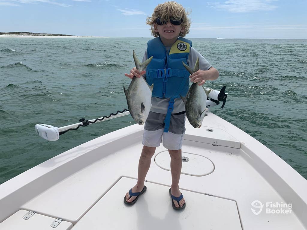 A young boy wearing a life jacket standing on the bow of a fishing boat with a trolling motor and the water behind him, holding two small Pompanos caught fishing in Florida