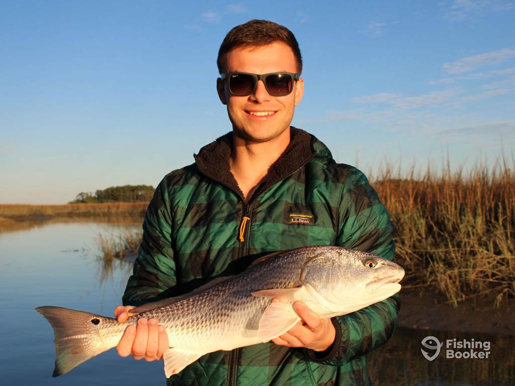 A smiling male angler in sunglasses, holds a Redfish with water and grassy flats behind him on a sunny afternoon