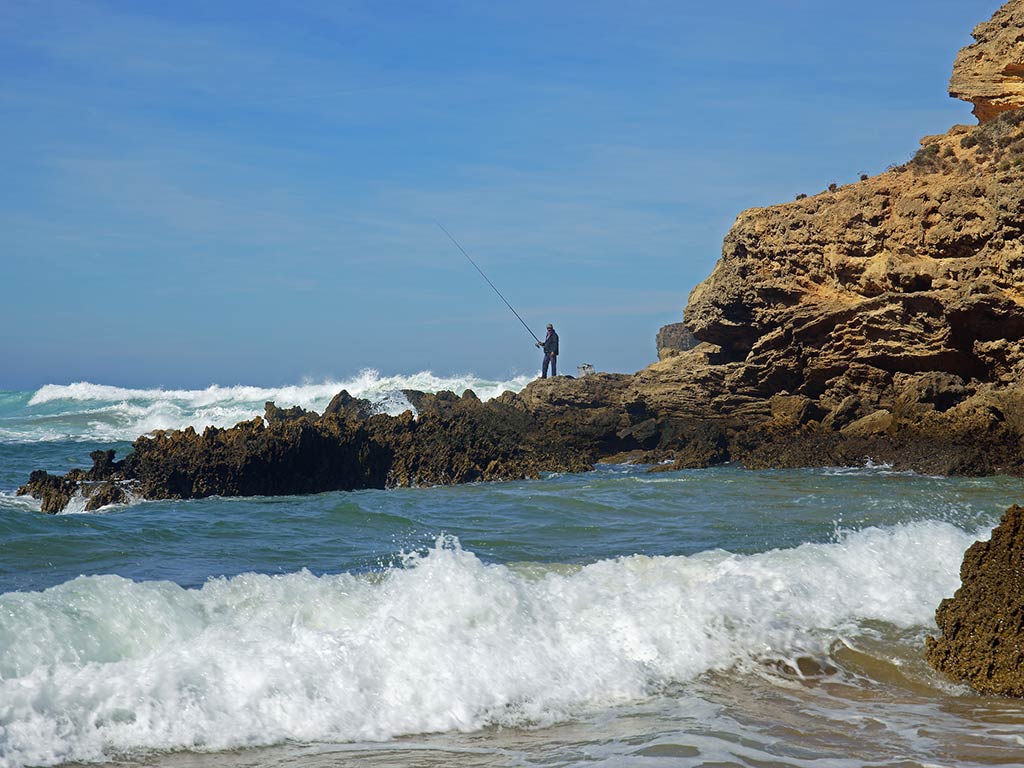 A view from a beach across crashing waves and a rocky shoreline towards a lone angler, casting a line in the Algarve