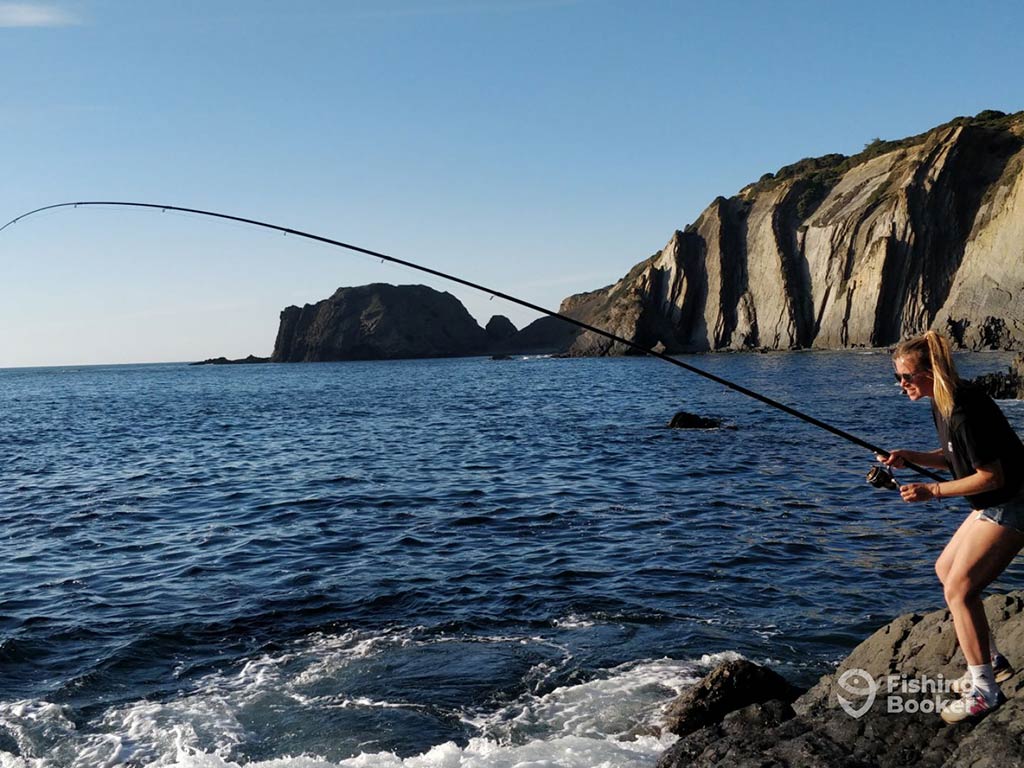 A woman casting a long line into the water from shore in the Algarve on a sunny day, with the rugged coastline and cliffs visible behind her