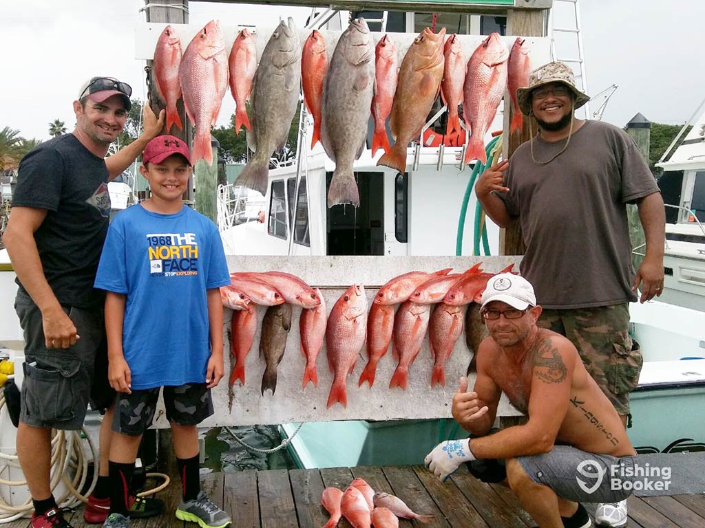 A group of anglers pose in front of a board back at the dock after a successful reef fishing trip in the Gulf of Mexico, showing off their Snapper and Grouper catches on a cloudy day