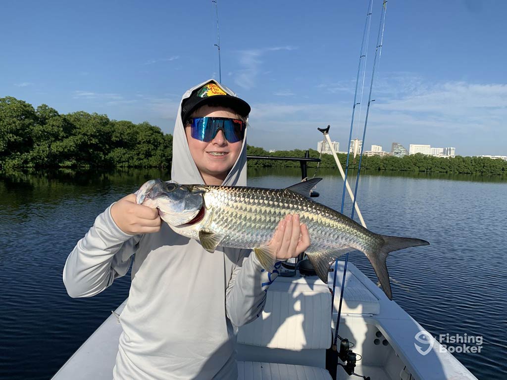 A youthful angler in sunglasses and with a hood over his baseball cap holds a Tarpon up to the camera, with water and mangroves behind him on a sunny day