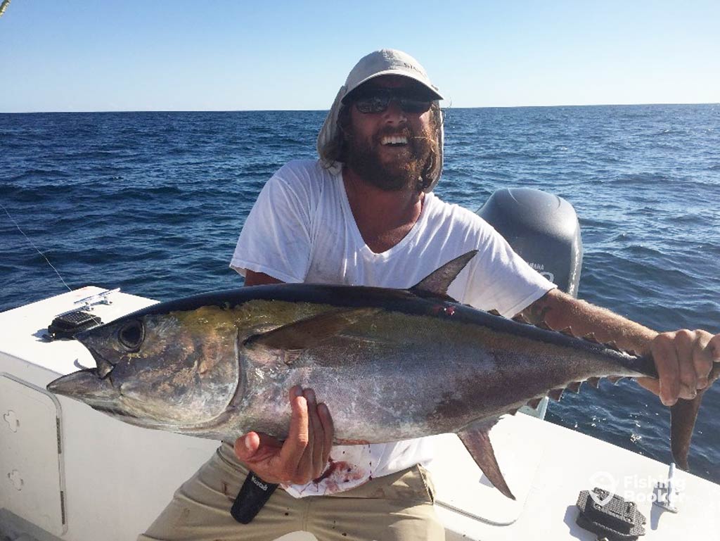 A smiling angler in a hat crouching down on a boat and holding a large Tuna with the water behind him on a sunny day