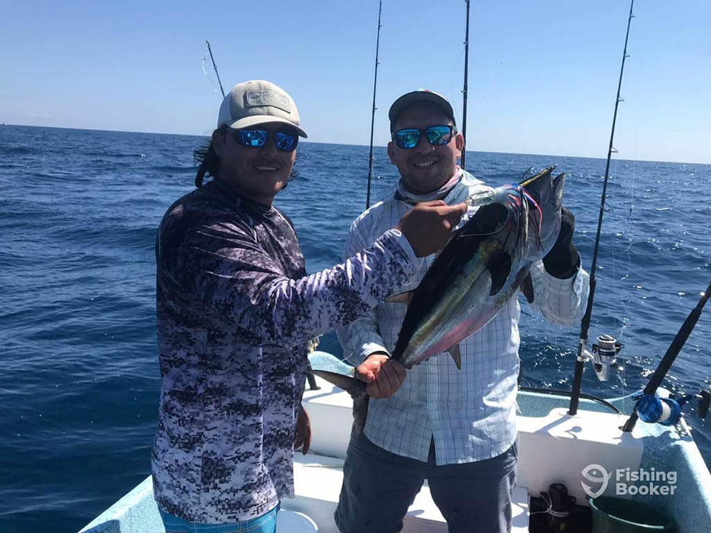 Two anglers in baseball caps and sunglasses hold a Tuna caught while offshore fishing in Puerto Morelos, while standing on a fishing charter with the water behind them on a clear day