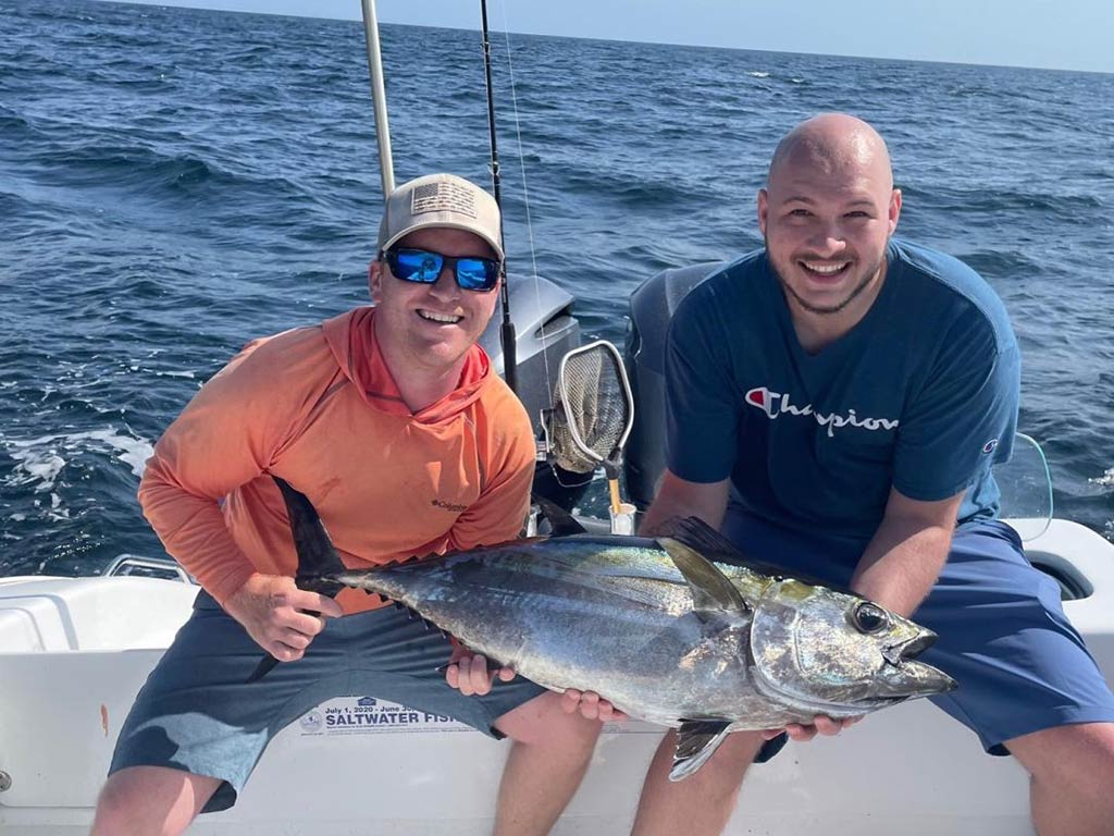 Two anglers crouching on the deck of a sportfishing boat, holding a Tuna caught offshore with the water behind them on a sunny day