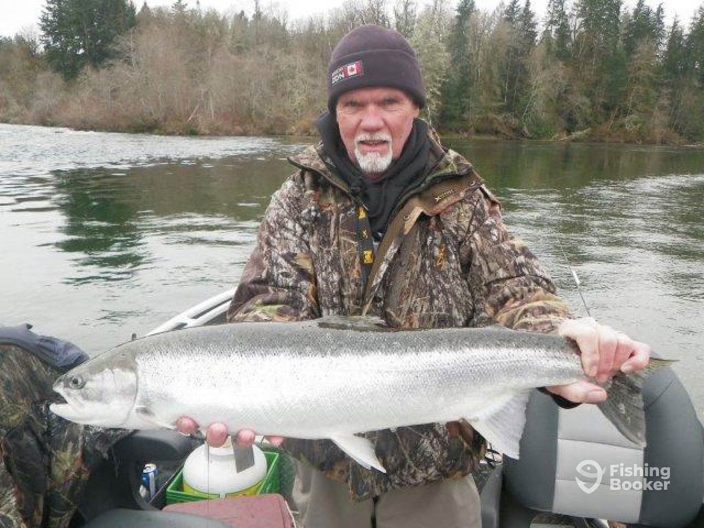 An elderly angler with a woolly hat and a grey beard holding a Sockeye Salmon on a boat on a river in Ucluelet, on a cloudy day