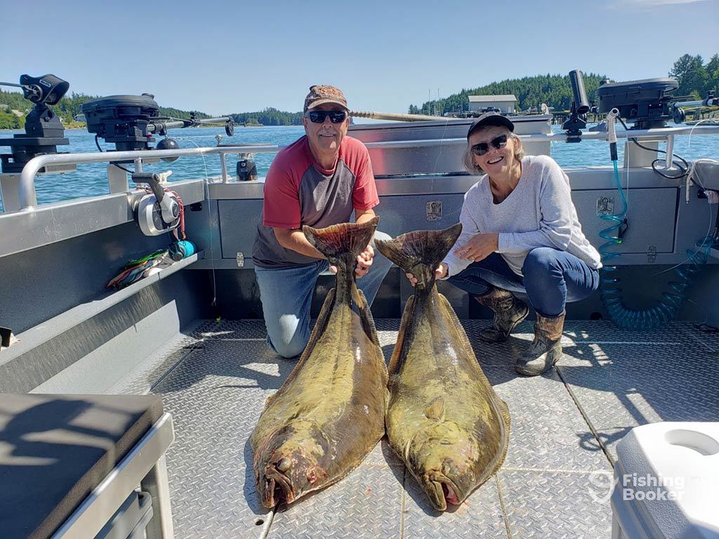 A man and a woman crouch behind two large Halibuts on a boat, caught while fishing in Ucluelet on a sunny day with the water behind them