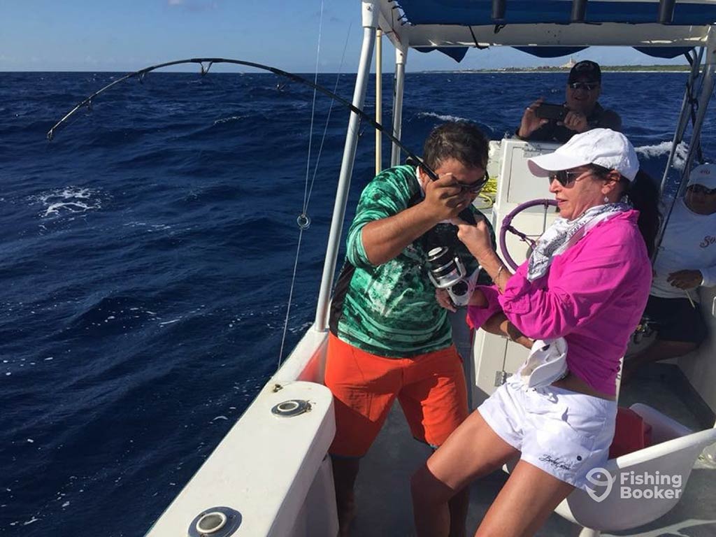 A man helps a woman battle a fish over the side of a boat, with dark blue waters visible on the left of the image on a sunny day