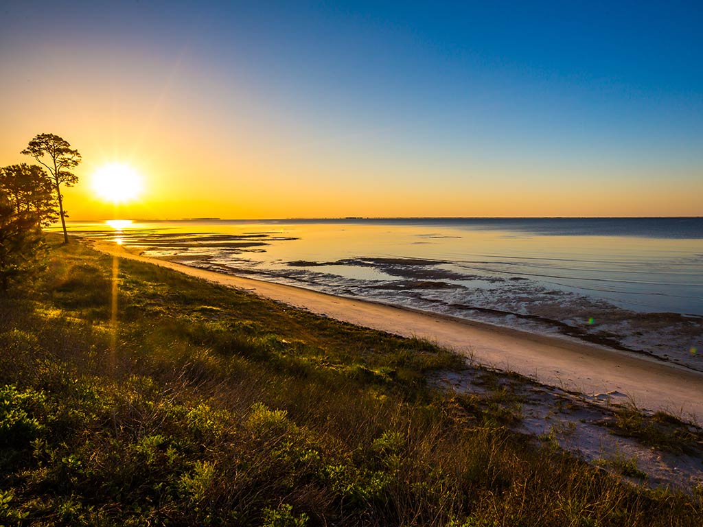 A view along a beach in Apalachicola at sunset with the sun visible in the distance on the left of the image and the sea on the right