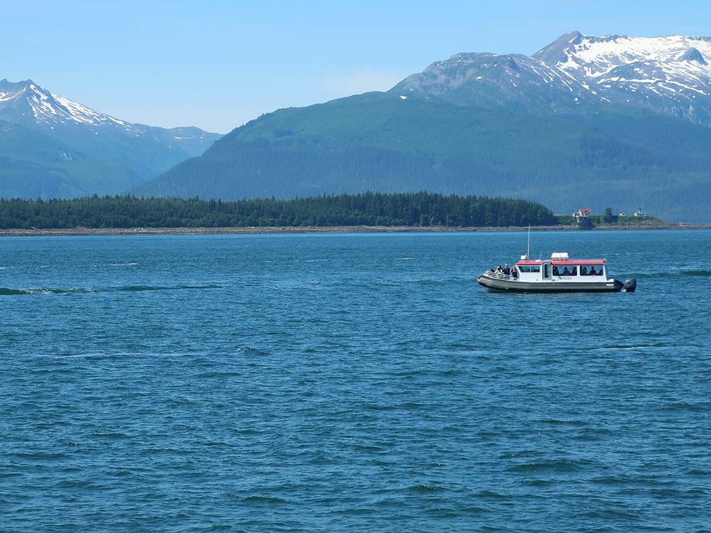 A view across the water towards a lone fishing boat on the waters of Juneau, AK, on a sunny day with snow-capped mountains in the distance