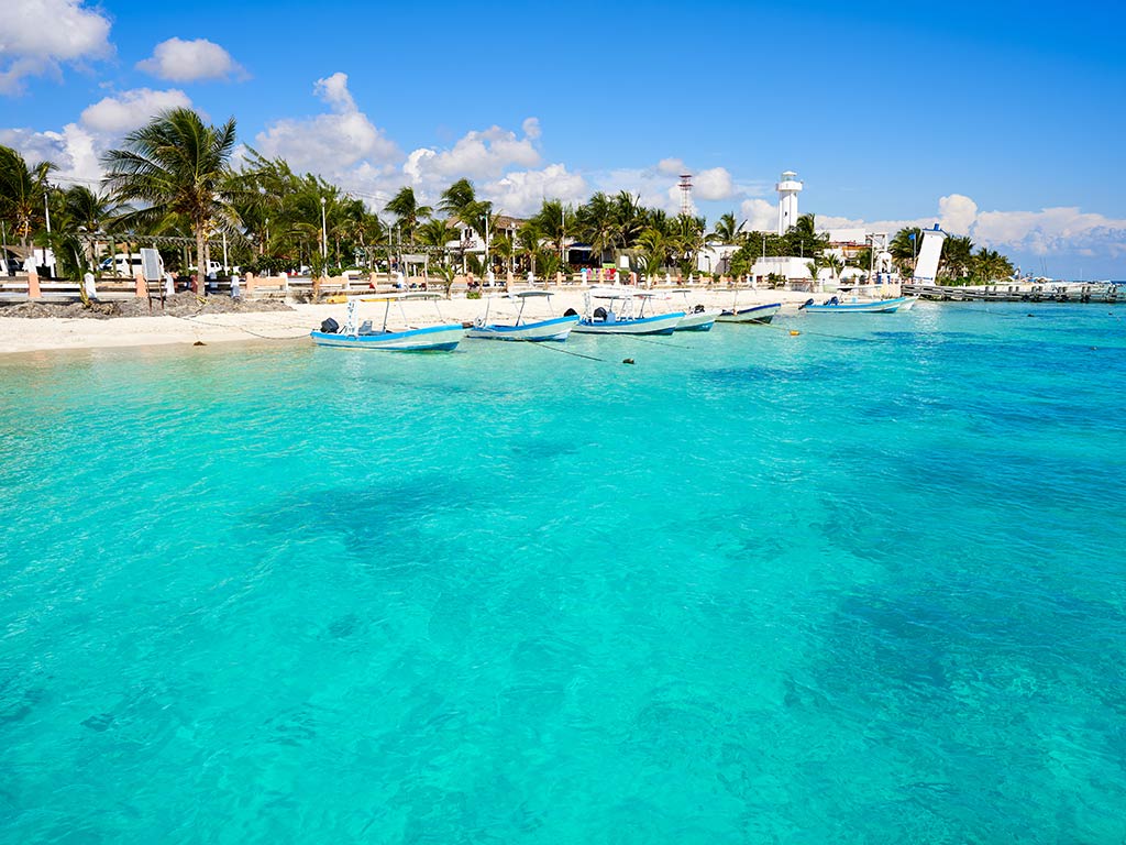 A view from the water to a beach in Puerto Morelos, with a number of small, traditional fishing boats docked in the sand, and trees lining the beach on a sunny day