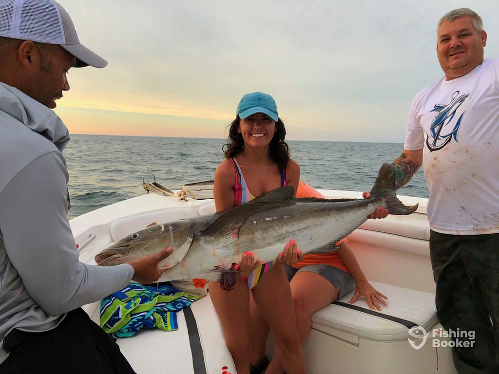 A female angler in a blue cap sitting on the bow of a fishing boat, holding a large Cobia on her lap, which is being supported by two men either side of the woman