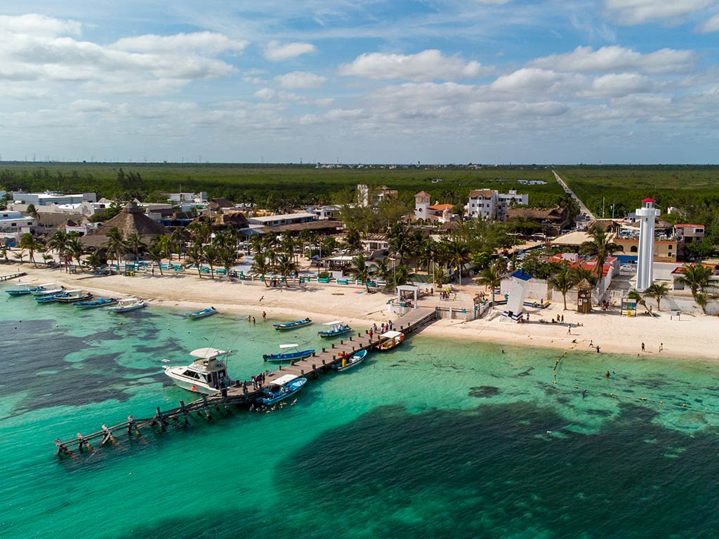 An aerial view of a fishing pier or small wooden boat dock in Puerto Morelos on a sunny day, with the turquoise waters of the sea visible in the foreground and the beach in the middle of the image